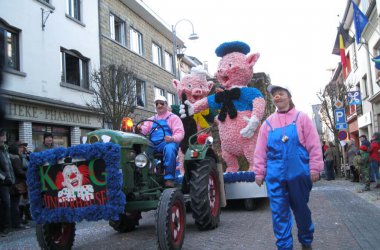 Traktor zieht Karnevalswagen mit rosa Schweinen in blauen Arbeiterhosen