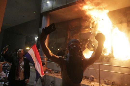 Mann mit Landesflagge steht vor brennendem Regierungsgebäude