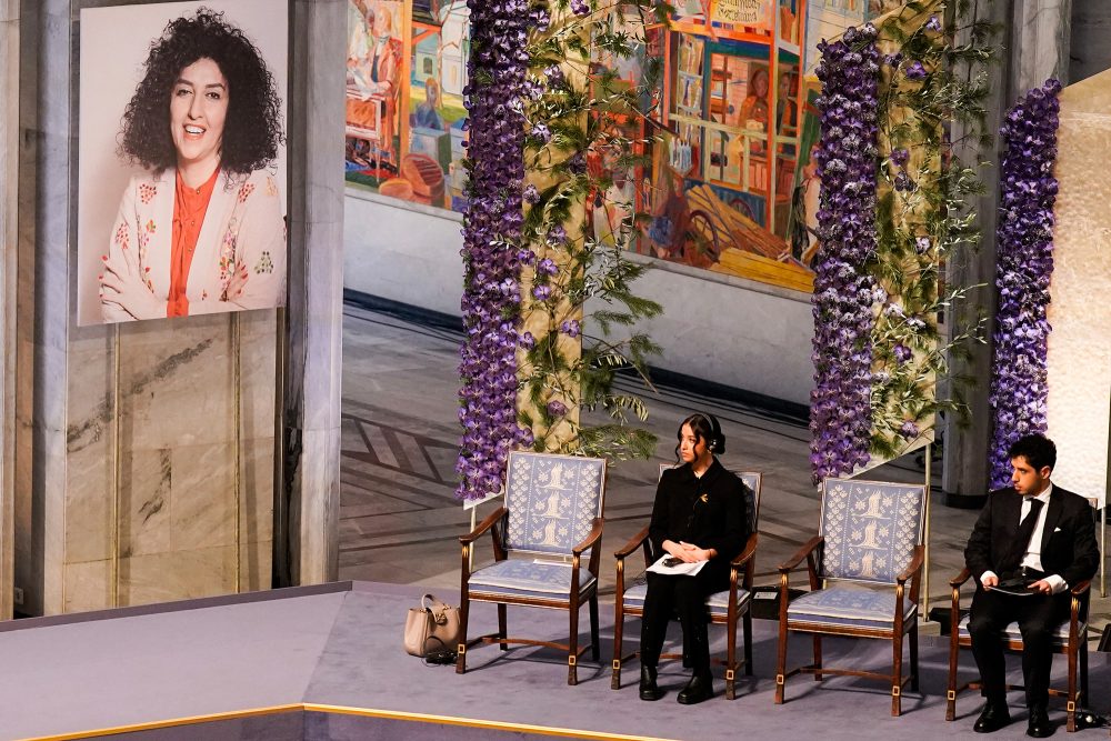 Ein Porträt der Friedensnobelpreisträgerin 2023 Narges Mohammadi hängt an der Wand, während ihre Kinder Kiana Rahmani und Ali Rahmani bei der Preisverleihung im Rathaus von Oslo neben einem leeren Stuhl sitzen (Bild: Fredrik Varfjell/NTB/AFP)