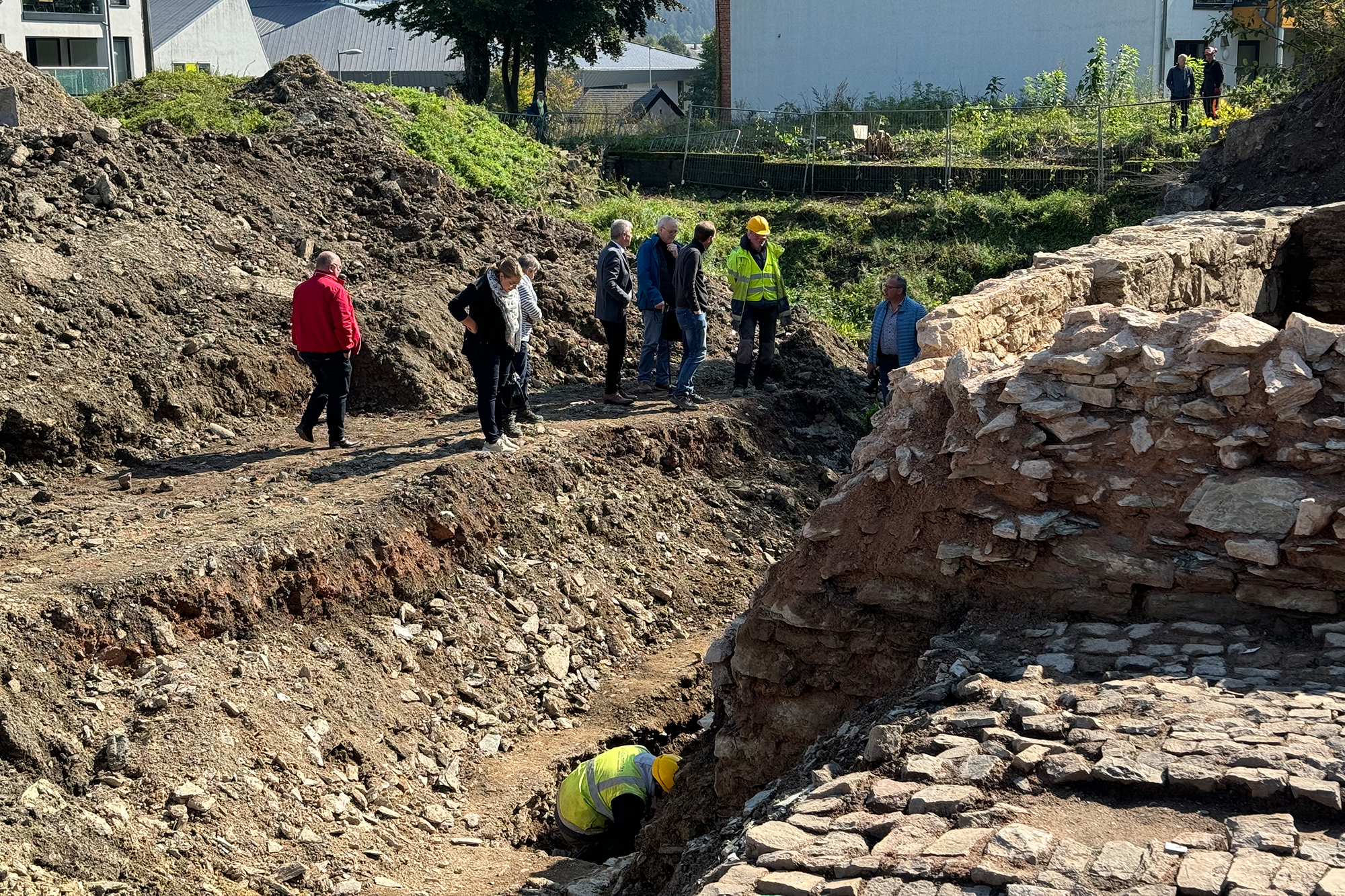 Ein Mann führt Ausgrabungsarbeiten an der Burg St. Vith durch, andere Menschen stehen in der Nähe und gucken ihm zu oder aber unterhalten sich