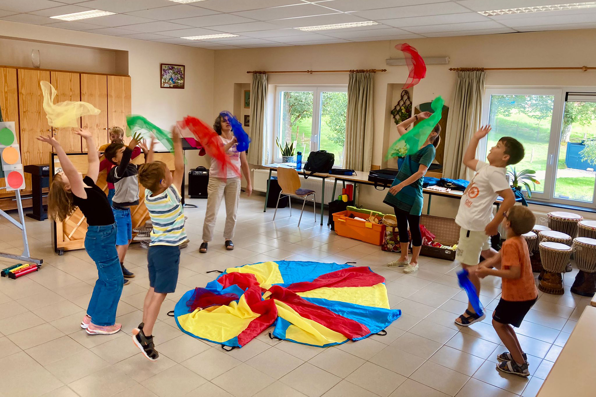 Kinder und eine Musikpädagogin bilden einen Kreis um ein kreisförmiges buntes Tuch auf dem Boden und werfen verschiedenfarbige kleine Tücher in die Luft