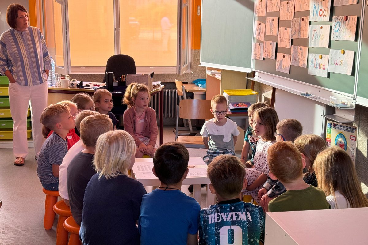 Die frisch gebackenen Primarschüler sitzen in ihrem Klassenraum im Stuhlkreis um einen kleinen Tisch herum, die Klassenlehrerin steht hinter ihnen