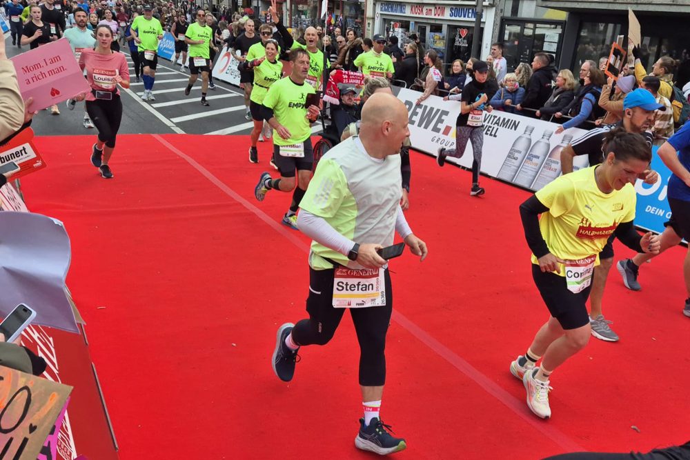 Läufer beim Halbmarathon im Köln, in hellgelben Trikots ist das Team Emdygo dabei, etwas versteckt mittig oben ist der Spezial-Rollstuhl