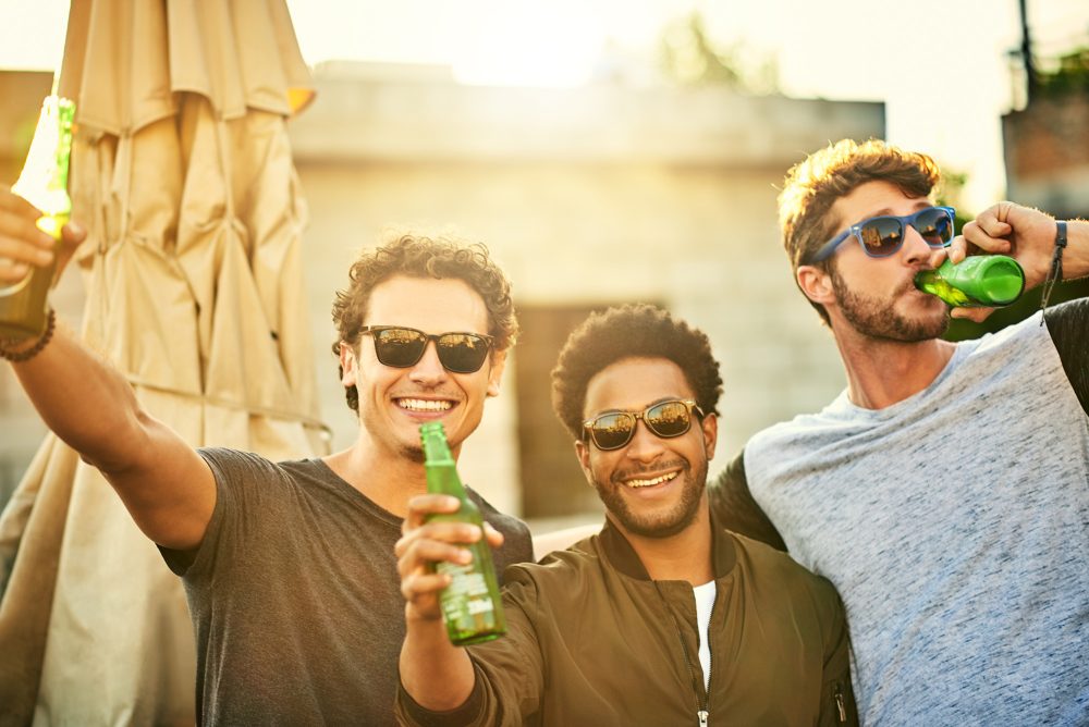 Junge Männer mit Bierflaschen in einer sommerlich anmutenden Feieratmosphäre lachen in die Kamera