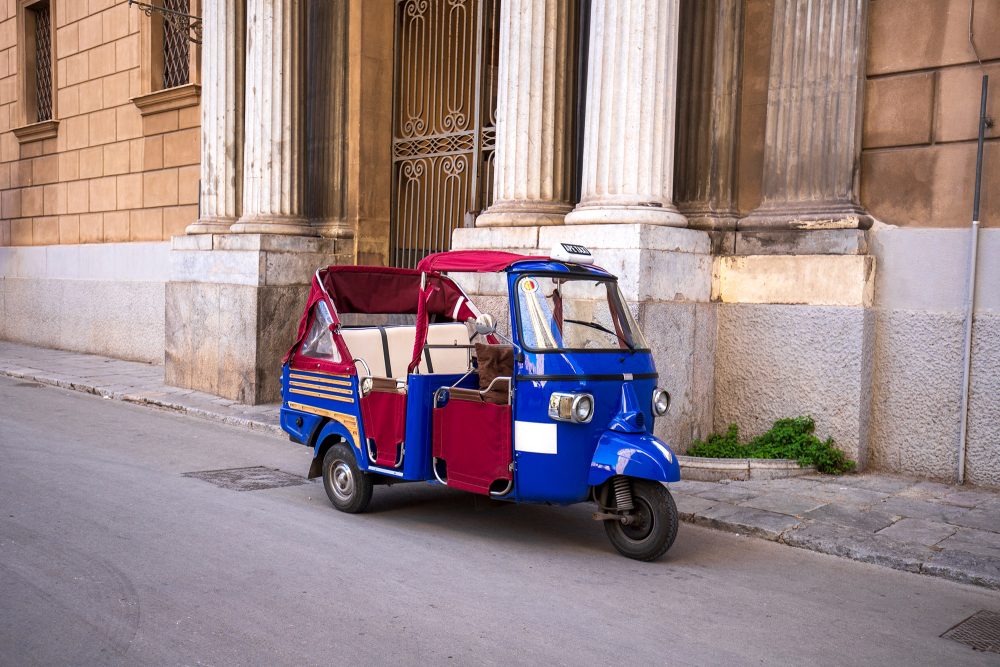 Der Kleintransporter "Ape" steht geparkt am Rand einer Straße in Italien, vor einem alten Gebäude mit antik anmutenden Säulen und einem großen eisernen Einfahrtstor