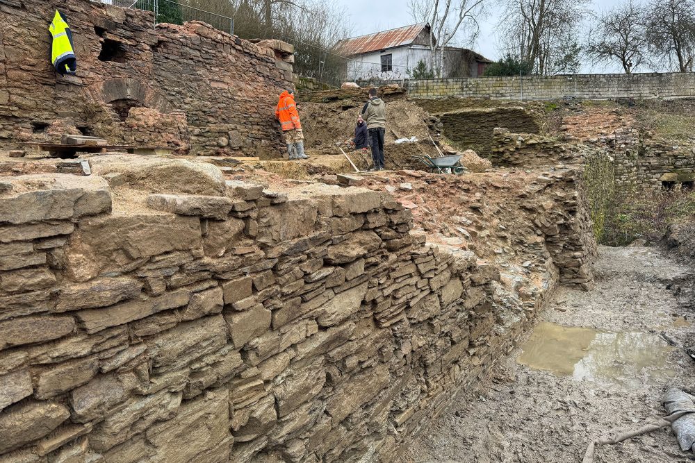 Drei Mitarbeiter stehen auf Mauerresten der Burg St. Vith und legen Steine frei