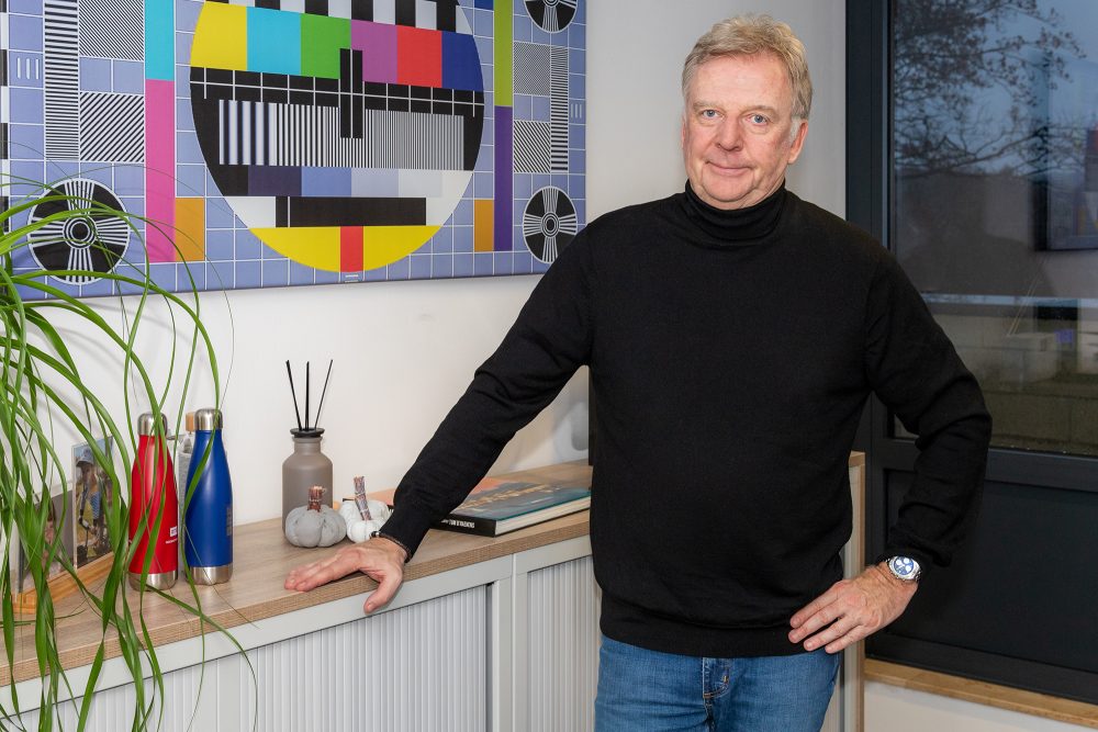 François de Brigode steht in einem Büro, im Hintergrund hängt an der Wand ein Bild mit dem früheren Fernseh-Testsignal-Motiv