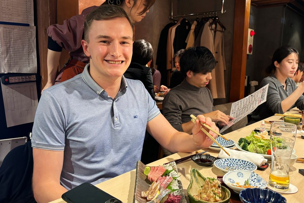 Maxim Luxen aus Medell sitzt am Tisch im Restaurant, in einer Hand hält er zwei Essstäbchen, auf dem Tisch stehen verschiedene Gerichte und ein Bierglas