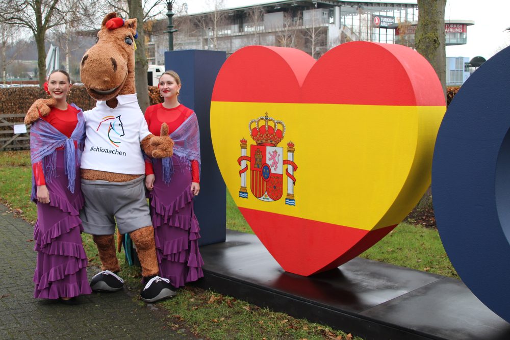 CHIO-Maskottchen mit zwei Flamenco-Tänzerinnen vor einer großen Herzskulptur in den spanischen Nationalfarben.