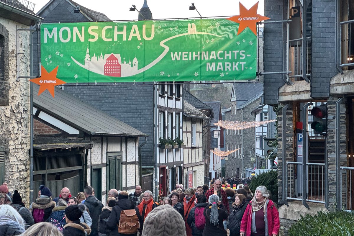 Menschen flanieren durch Monschau, zwischen den Häusern hängt ein befestigtes Banner mit der Aufschrift "Monschau - Weihnachtsmarkt"