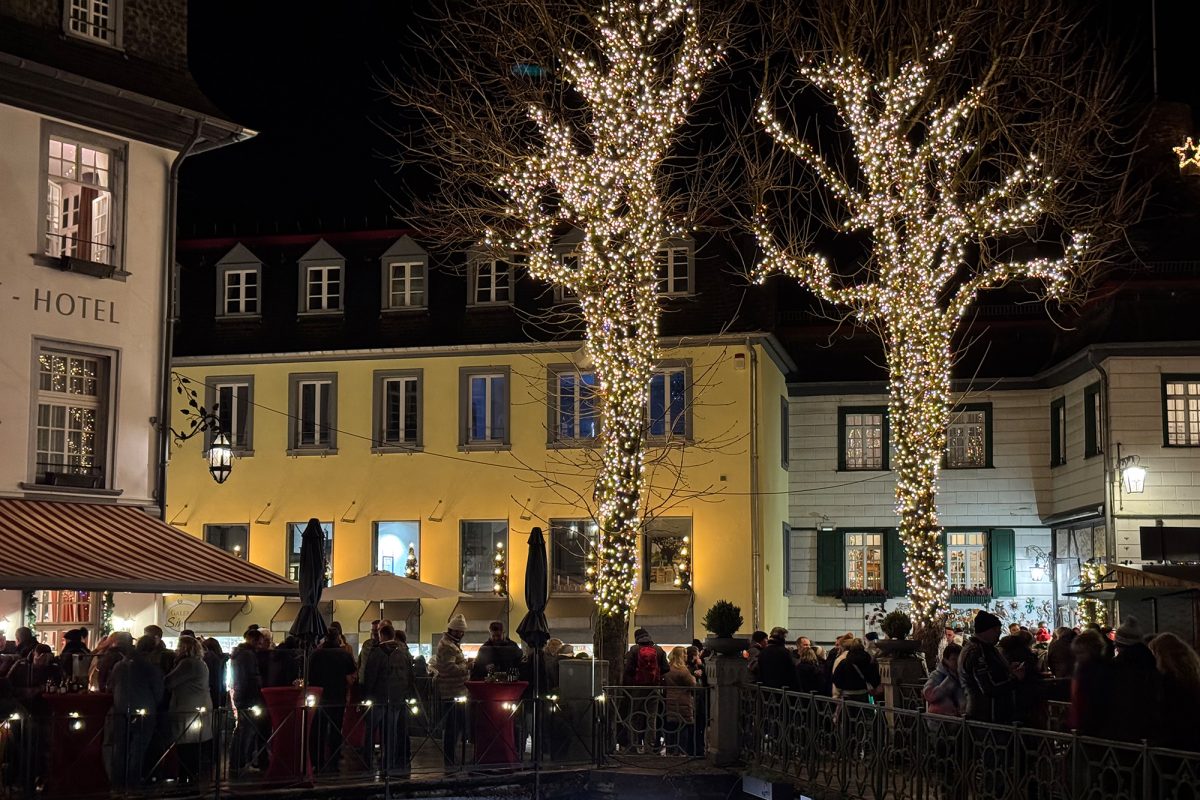 Zwei durch Lichterketten illuminierte Bäume auf einem Platz in Monschau, auf dem viele Menschen stehen