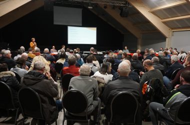 Der Saal ist voller Menschen, die hier von hinten auf Stühlen sitzend zu sehen sind - weiter vorne ist die Bühne, auf der eine Leinwand steht, auf die etwas projiziert wird