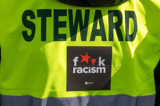 Auf der Warnweste eines Ordners mit der Aufschrift "Steward" klebt ein Aufkleber mit der Aufschrift "Fuck Racism"