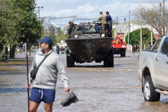 Ein gepanzertes Armeefahrzeug fährt durch eine überflutete Straße, im Vordergrund watet ein Mann durch das Wasser und hält einen Schuh in der Hand