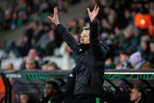 Trainer Jimmy De Wulf steht neben dem Fußballfeld und hält beide Hände in die Höhe