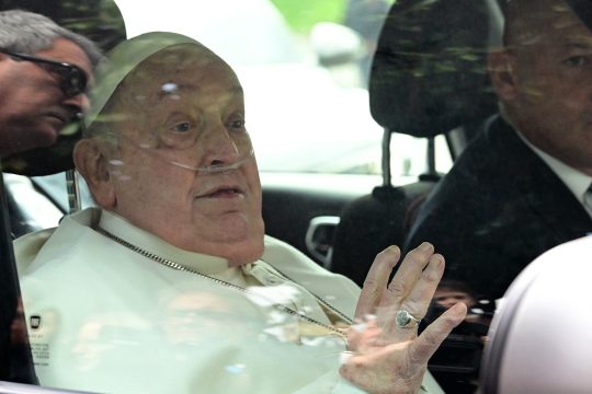 Papst Franziskus sitzt in einem Wagen, er hat eine Atemhilfe an der Nase, schaut aus dem Autofenster und hält eine Hand leicht hoch