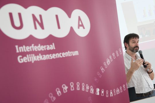 Unia-Co-Direktor Patrick Charlier hält ein Mikrofon und spricht auf einer Pressekonferenz - den meisten Teil des Fotos macht ein Banner von Unia aus