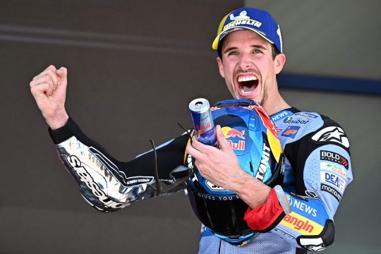 Alex Marquez reckt jubelnd einen Arm in die Höhe, unter dem anderen Arm klemmt sein Helm und in der Hand hält er eine Getränkedose