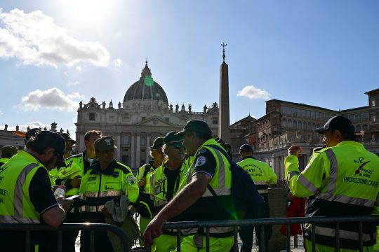 Mitglieder des Zivilschutzes stehen neben einer Absperrung mit dem Petersdom und dem Obelisken des Vatikans im Hintergrund