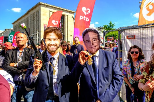 Zwei Demonstranten haben Masken mit den Gesichtern von George-Louis Bouchez und Bart De Wever auf, Bouchez hält eine Gewehrattrappe in die Luft, aus deren Lauf eine Plastikblume schaut