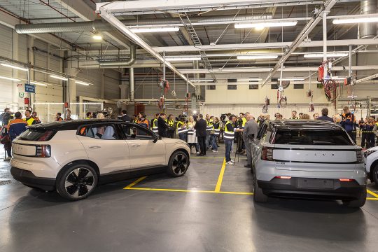 Zwei Fahrzeuge des Modells EX30 stehen in einer Werkshalle, im Hintergrund stehen viele Mitarbeiter