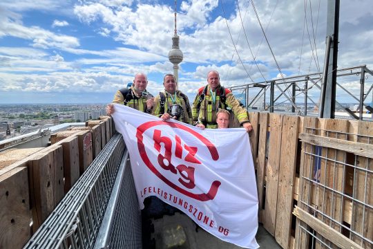 René Schoonbrood, Jörg Heinen, Daniel Klinges und Leif Daniels stehen auf dem Dach und halten die Flagge der Hilfeleistungszone DG in den Händen, im Hintergrund sieht man den Berliner Fernsehturm