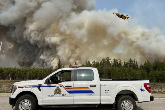 Im Vordergrund ein Polizeiwagen, im Hintergrund ein Waldbrand mit großer Rauchwolke und einem Löschflugzeug am Himmel