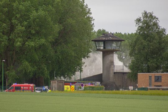 Rauchwolke über dem Gefängnisgelände, nahe der Außenmauern mit Wachturm sind Feuerwehrfahrzeuge zu sehen