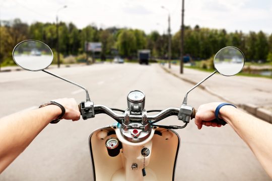 Person fährt auf Motorroller eine Straße entlang, aus der Ich-Perspektive sieht man zwei Arme, die den Lenker festhalten