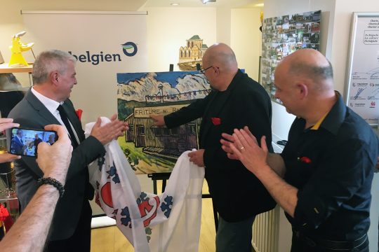 Künstler Eric Legrain (Mitte) erklärt etwas zu dem Bild, das er extra für den Einzug der ostbelgischen Fritte ins Micro-Frittenmuseum in Forest gemalt hat