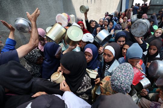 Bedürftige Palästinenserinnen stehen mit Gefäßen Schlange an einer Essensausgabe im Gazastreifen