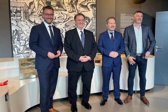 Der Kelmiser Bürgermeister Daniel Hilligsmann, Botschafter Martin Kotthaus, der Föderalabgeordnete Luc Frank und der EU-Abgeordnete Pascal Arimont stehen in einem Raum des Museum Vieille Montagne