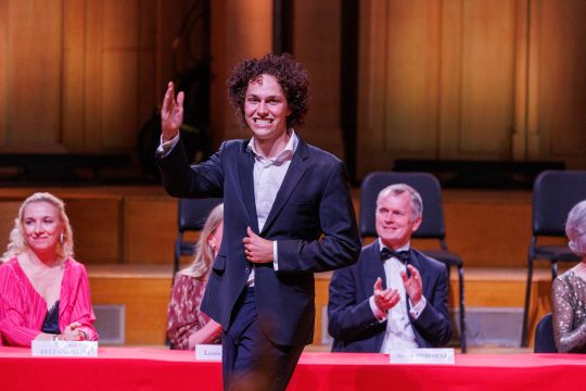 Der niederländische Pianist Nikola Meeuwsen schaut sichtlich erfreut und winkt mit einer Hand, hinter ihm sitzen Jurymitglieder an einem Tisch