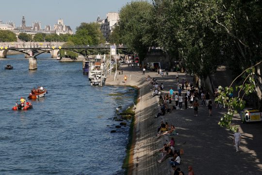Am Seineufer in Paris spazieren Menschen entlang, einige sitzen auf dem Boden direkt neben dem Wasser