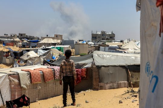 Kind in einem Flüchtlingslager in Rafah - in der Ferne sieht man Rauch aufsteigen