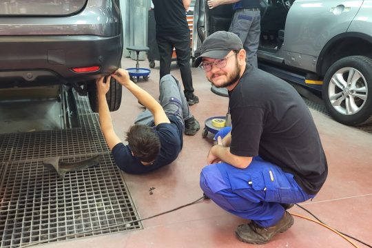 Ein Mitarbeiter liegt rücklings auf dem Boden und führt Arbeiten an einem Fahrzeug aus, Ben Olbertz rechts im Bild hockt daneben und lächelt