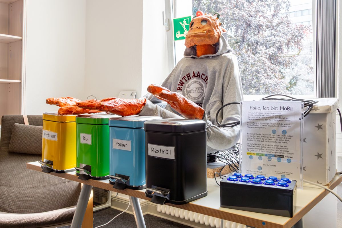 Roboter mit monsterähnlichem Gesicht und mehreren Armen, an deren Enden riesige Hände zu sehen sind - auf einem Tisch stehen vier verschiedenfarbige Mülleimer für Wertstoffe, Bio, Papier und Restmüll. Auf einem Erklärungsschild steht etwas über den Roboter, der "MoBi" heißt.