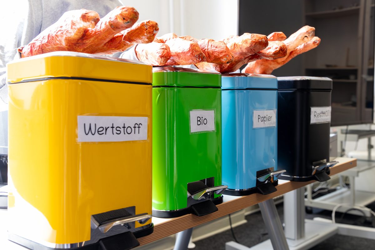 Vier unterschiedlich farbige Mülleimer für unterschiedliche Müllsorten, auf ihnen liegen Roboterbestandteile, die an mehrere riesige monsterähnliche Hände erinnern