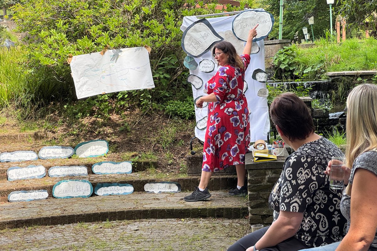 Elke Drosson zeigt auf ein Kärtchen mit der Aufschrift "Ich wohne", das auf einem Whiteboard aufgeklebt ist, welches im Garten steht - daneben gibt es noch andere Kärtchen mit Begriffen und kurzen Sätzen