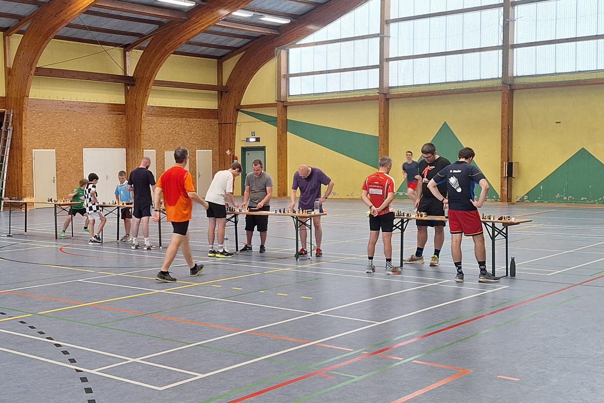 In der Sporthalle sind mehrere Tische mit Schachbrettern aufgebaut, um sie herum stehen die Spieler