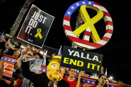 Demonstranten in Tel Aviv - einer hat eine Maske an, die Donald Trumps Gesicht darstellen soll - außerdem werden Schilder hochgehalten - auf einem steht "Yalla. End it!", auf dem anderen "Just do it"