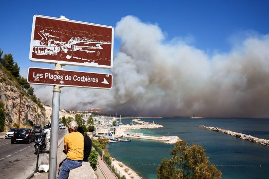 Menschen beobachten in der Nähe der Plage des Corbieres in Marseille, wie im Hintergrund Rauch von einem Waldbrand aufsteigt
