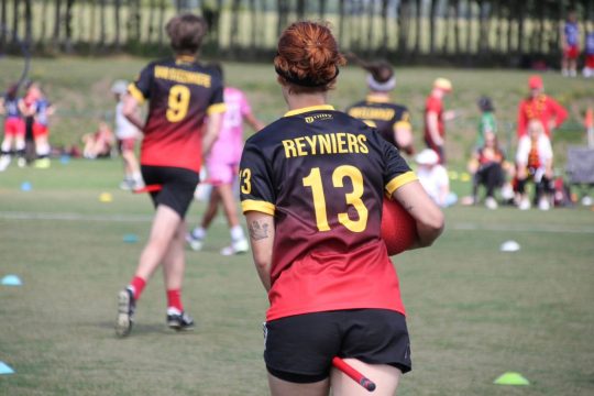 Quadball-Spielerinnen und Spieler von hinten, bei der Person vorne im Bild sieht man das Plastikrohr zwischen den Beinen