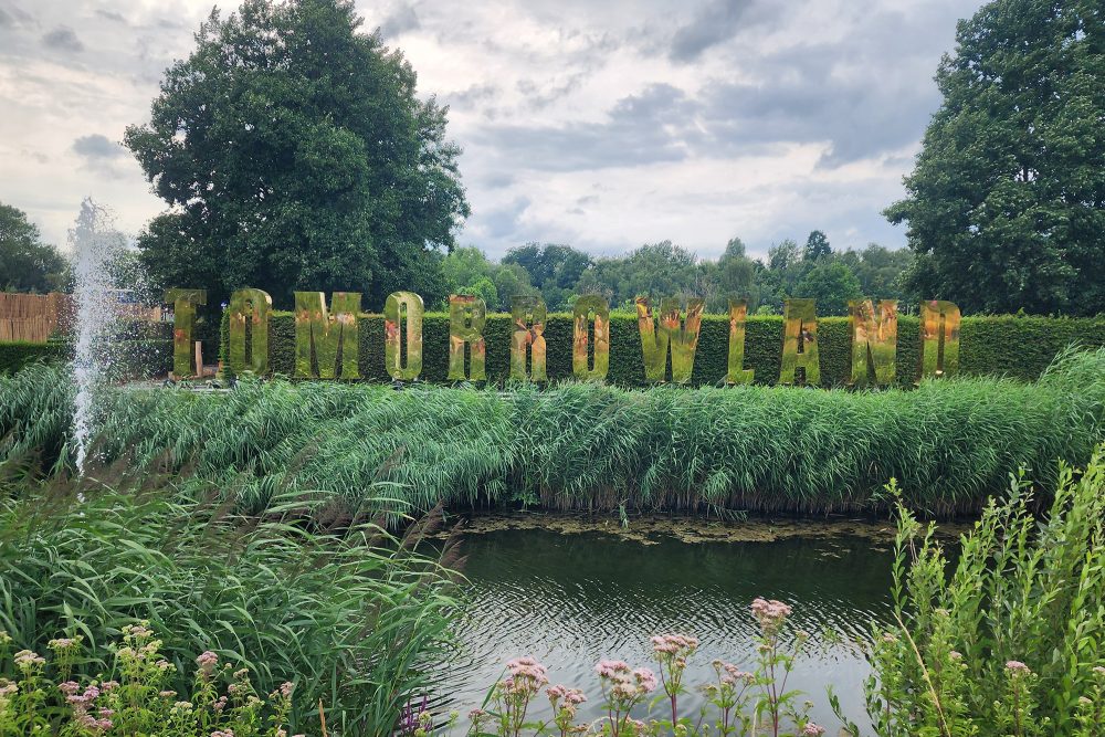 Schriftzug von Tomorrowland in großen goldenen Buchstaben vor einem kleinen See mit Springbrunnen