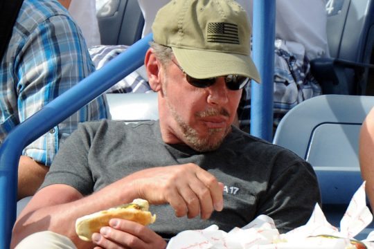 Oracle-CEO Larry Ellison hat einen Hotdog in der Hand und sitzt in einem Stadion