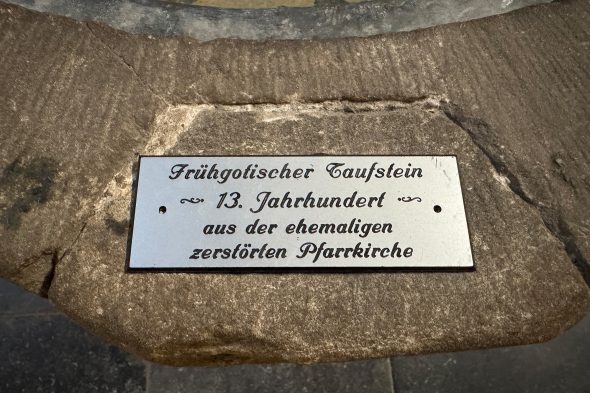 Nahaufnahme eines Schilds mit der Aufschrift: Frühgotischer Taufstein, 13. Jahrhundert, aus der ehemaligen zerstörten Pfarrkirche