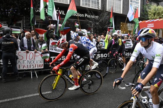 Beim Etappenstart in O Barco de Valdeorras halten Demonstranten hinter der Absperrung palästinensische Flaggen in die Höhe