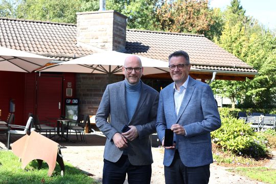 Städteregionsrat Dr. Tim Grüttemeier und Roetgens Bürgermeister Jorma Klauss posieren mit Smartphones in den Händen für ein Foto