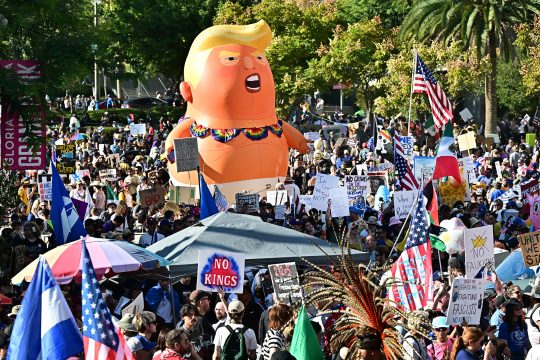 Demonstranten mit Transparenten und Plakaten, inmitten von ihnen eine riesige aufgeblasene Figur, die Donald Trump darstellen soll