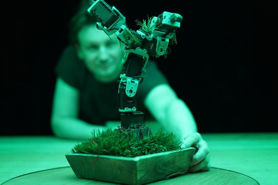 In grünem Lichtschein steht eine Figur auf einem Tisch, dahinter ein Mann